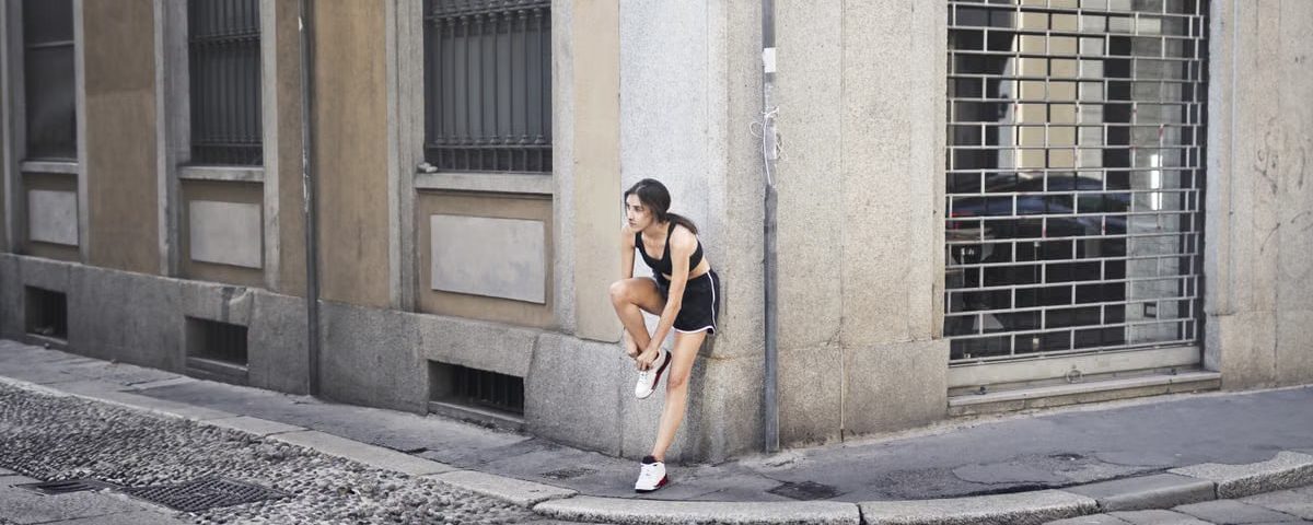 Slim female runner in activewear standing on corner of street and tying shoelaces on sneakers while leaning on stone wall of building and looking away