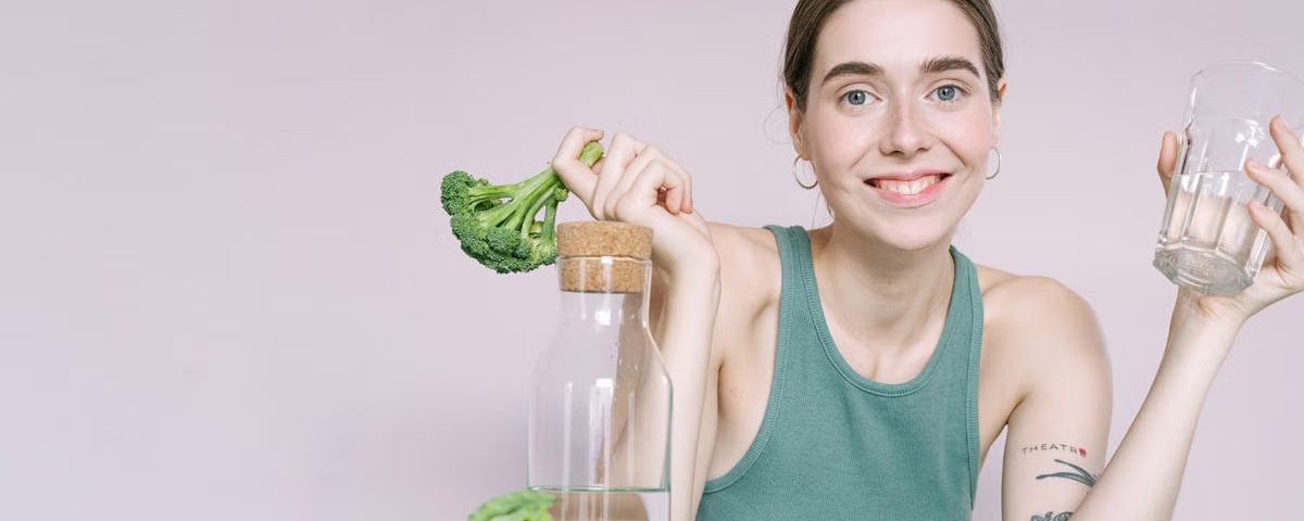 Woman in tank top smiling with broccoli, cabbage, and glass of water on table.